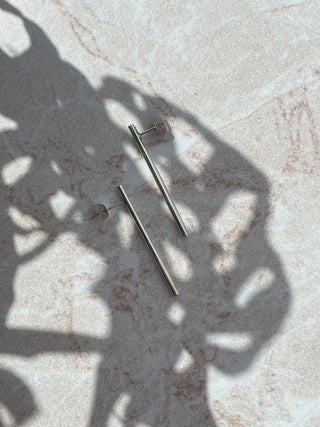 A pair of minimalist silver bar stud earrings with butterfly backings, displayed on a light beige textured surface with artistic leaf shadows.
