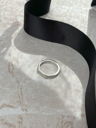 A silver flat top ring displayed on a textured light gray surface with a black ribbon draped behind it.