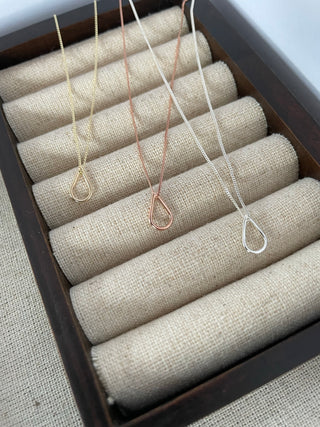 Three delicate teardrop pendant necklaces in gold, rose gold, and silver, displayed on a dark wooden jewelry organizer with beige fabric rolls.