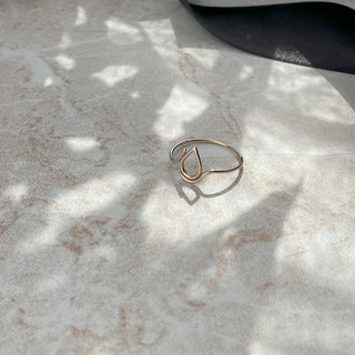 A delicate gold wire ring with a teardrop-shaped design, resting on a light marble surface with dappled sunlight and shadows. A black ribbon and a white ceramic bowl are blurred in the background.