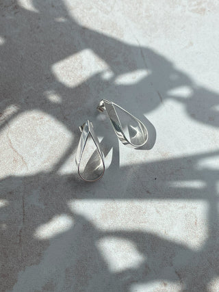 A pair of silver teardrop-shaped hoop earrings with a twisted, open design, resting on a light gray textured surface under dappled sunlight and shadows.