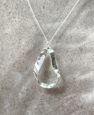 A recycled silver teardrop-shaped statement pendant necklace with a textured, hammered finish, hanging from a delicate silver chain against a neutral, light gray stone background.