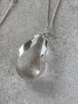 A polished recycled silver teardrop-shaped sculptural statement pendant necklace with a twisted, open design, resting on a textured gray stone surface.