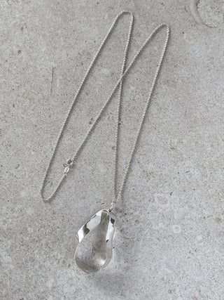 A recycled silver pendant necklace with a polished, sculptural and organic teardrop-shape on a delicate chain, displayed on a textured light gray stone surface.