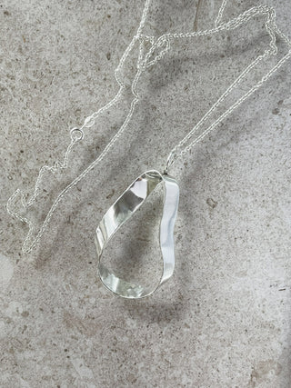 A recycled silver teardrop-shaped pendant necklace with a hammered texture, resting on a textured gray stone surface.