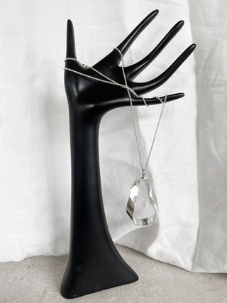 A recycled silver abstract sculptural statement pendant necklace displayed on a black hand-shaped jewelry stand against a white draped fabric background.