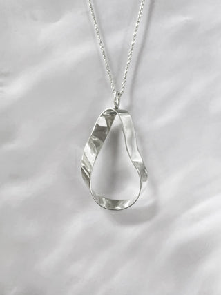 A silver teardrop-shaped pendant necklace with a textured, hammered finish, hanging from a delicate silver chain against a white fabric background.