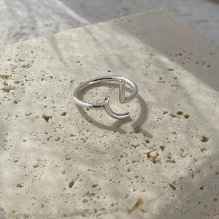 A silver open-band ring with a small curved hook detail, resting on a textured beige stone tile with soft natural shadows.