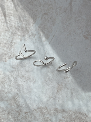 Three minimalist silver rings with abstract curved and infinity-like designs, arranged on a textured light gray surface with soft natural shadows.