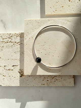 A thin silver-toned bangle bracelet with a small black bead, displayed on a textured beige stone tile under direct sunlight.