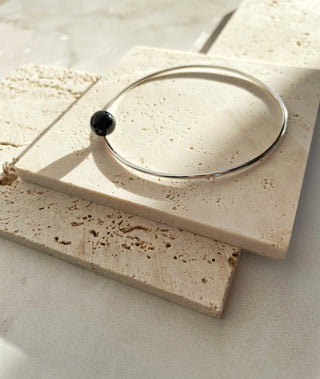 A thin silver-toned bangle bracelet with a small black spherical bead accent, displayed on a textured beige stone tile.