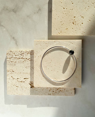 A thin silver-toned huggie bangle bracelet with a small black bead accent, displayed on a textured beige stone tile over a marble surface.