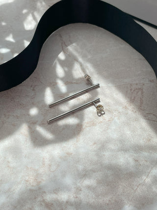 A pair of silver barbell stud earrings with small clear crystal accents, displayed on a light marble surface with a black ribbon prop and dappled sunlight.