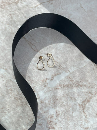 A pair of gold teardrop hoop earrings resting on a light beige marble surface, with a black ribbon draped behind them.